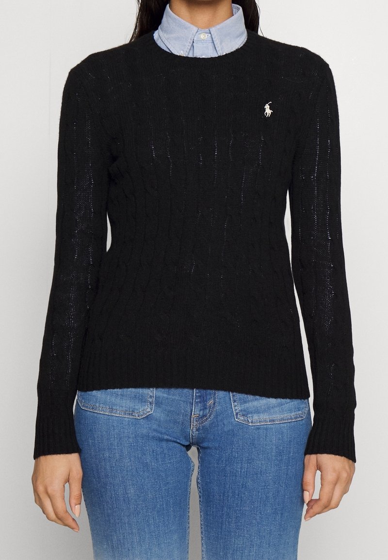 Personne portant un pull en maille torsadée noir par-dessus une chemise à col bleu clair, associée à un jean en denim bleu, debout devant un fond uni.