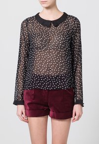 Blusa negra transparente con lunares blancos, cuello redondeado, mangas largas y puños, combinada con pantalones cortos de terciopelo burdeos.
