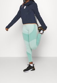 Persona che si allunga una gamba dietro, indossando un felpa blu navy, leggings leggeri a fantasia blu chiaro e sneakers Puma nere e bianche, su uno sfondo semplice.