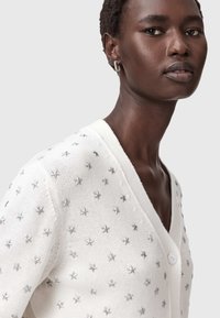 Weißer gestrickter Cardigan mit silbernen Sternapplikationen, V-Ausschnitt und einem einzigen Knopfverschluss. Weicher Stoff mit legerer Passform.