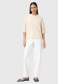 Maglione color crema con maniche corte, orlo a coste e dettagli delle cuciture verticali. Indossato con pantaloni bianchi a gamba larga e scarpe nere flat.