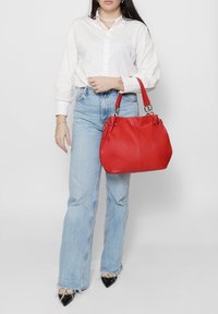 Borsa in pelle rossa con forma arrotondata, ferrature argentate e doppi manici. Il modello indossa una camicia bianca con bottoni e jeans azzurri chiari.