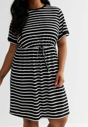 Kvinde iført en sort og hvid kortærmet kjole med vandrette striber og snøre i taljen, stående foran en ensfarvet baggrund.