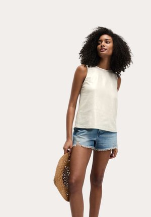 Lys beige ærmeløs top i en glat tekstur, parret med slidte denimshorts og holder en vævet stråhat.