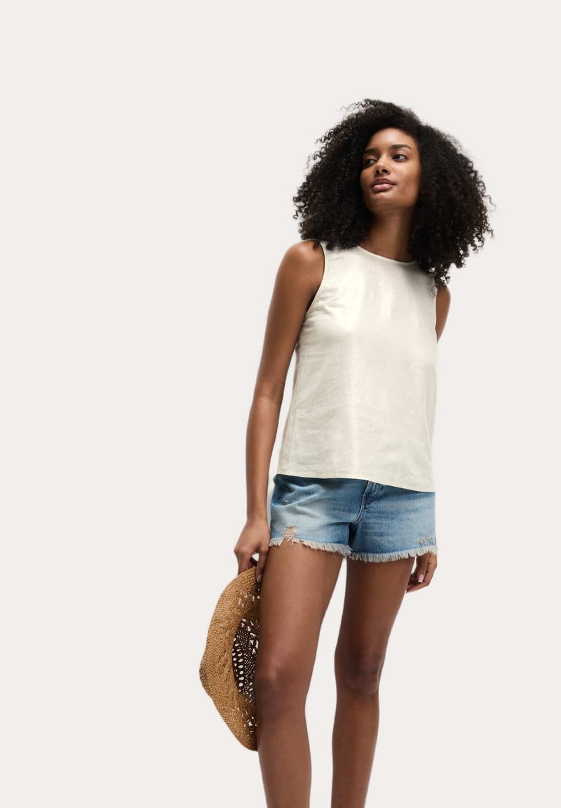 Lichtbeige mouwloze top in een soepele textuur, gecombineerd met versleten denim shorts, en houdt een geweven strooien hoed vast.