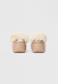 Beige slip-on klumper med myk faux pels trim og teksturert gummisåle, med en glatt læraktig kropp og avrundet design.
