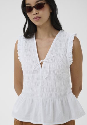 Woman wearing dark sunglasses and a white sleeveless blouse with ruffled shoulders and a tie at the neckline, set against a plain background.