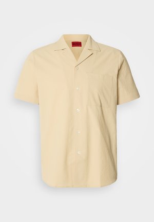 Chemise à manches courtes, de couleur jaune clair, en tissu texturé. Elle présente un col pointu et une poche poitrine, avec des boutons blancs.