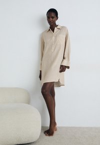 Cremefarbenes, langärmliges Kleid mit geometrischem Muster, Knopfkrause, seitlichen Schlitzern und weicher Textur, kombiniert mit baren Füßen.