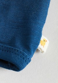Manche en tissu de coton bleu avec une texture côtelée et une étiquette crème ornée d'un petit canard jaune brodé. Couture lisse le long du bord.