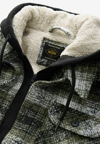 Fleeceforet jakke med lynlås i et ternet mønster i grøn og sort, med en sort krave og holdbart hardware samt en label indeni.