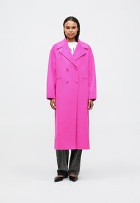 Sieviete stāv, vērsta uz priekšu, ģērbta garā spilgti rozā divrindu mētelī, melnās biksēs un melnās smailējošās kurpēs uz balta fona.