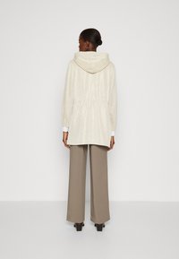 Giacca leggera con cappuccio, vita elastica, polsini a coste e finitura testurizzata. Abbinata a ampi pantaloni beige e tacchi a blocco.
