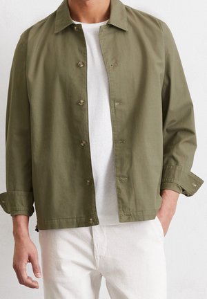 Mann trägt eine olivgrüne Jacke mit Knöpfen über einem weißen Hemd und weißen Hosen, steht mit einer Hand in der Tasche.