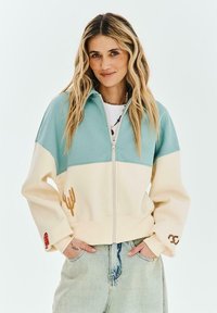Žena s dlouhými blond vlasy nosící bundu na zip ve světle modré a krémové barvě, světlé džíny a náhrdelník z korálků, ruce v kapsách.