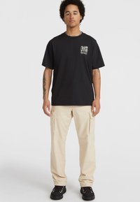 Sort bomulds T-shirt med et grafik på venstre bryst, parret med beige cargo bukser med sidelommer og sort fodtøj.