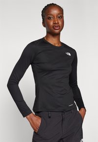 Maglietta nera a maniche lunghe con scollo tondo, caratterizzata da un tessuto strutturato e un logo bianco sul petto sinistro, abbinata a pantaloni scuri.