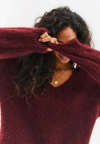 Burgundowy sweter z głębokim dekoltem w serek, o fakturowanym splocie i długich rękawach. Materiał wydaje się miękki i puszysty, co podkreśla jego przytulny wygląd.