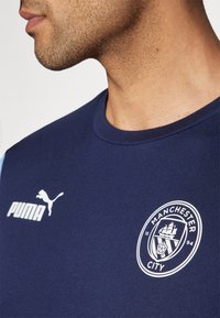 Námořnická modrá sportovní tričko vyrobené z bavlny, s bílým logem Puma na levém hrudníku a emblémem Manchester City na pravé straně.