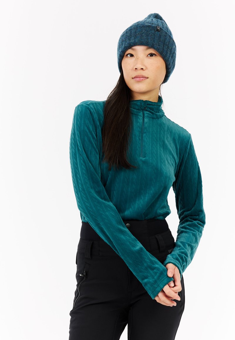 Turkusowa bluzka z długim rękawem, z połową zamka błyskawicznego przy kołnierzy i teksturowanym wzorem, zestawiona z czarnymi spodniami z zamkami błyskawicznymi w kieszeniach i dopasowanym pasem.