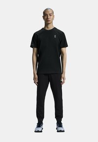 T-shirt noir à manches courtes avec un col rond et un logo sur la poitrine, associé à un pantalon noir ajusté et des baskets grises.