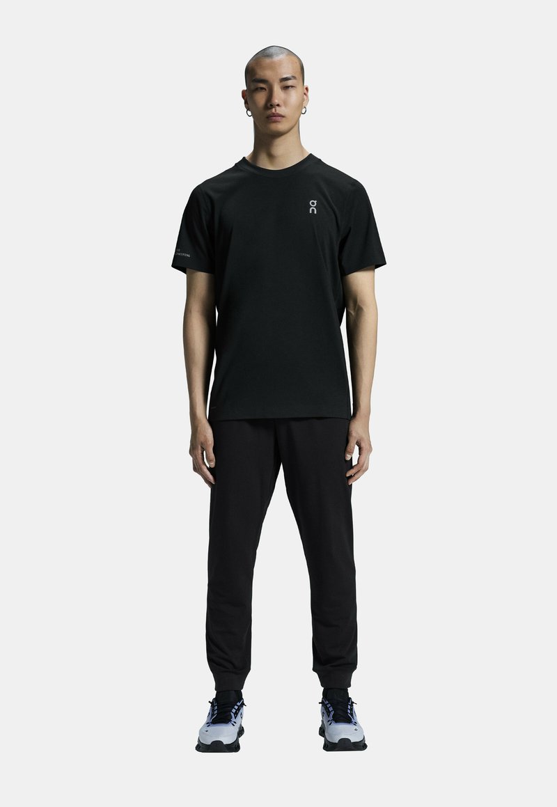 T-shirt noir à manches courtes avec un col rond et un logo sur la poitrine, associé à un pantalon noir ajusté et des baskets grises.