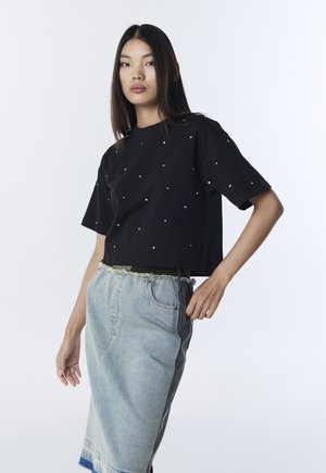 Junge Frau trägt ein kurzes schwarzes T-Shirt mit kleinen weißen Formen und einen zweifarbigen Jeansrock mit ausgefranstem Saum, posiert vor weißem Hintergrund.