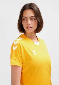 Gul sportströja av lätt tyg. Har vit logotyp och axelaccents. Kort ärm med avslappnad passform och v-ringning.