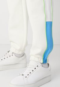 Hvide svedbukser med blå og neon grønne sideaccentuer, der har elastiske manchetter. Kombineret med hvide sneakers og grå sokker.