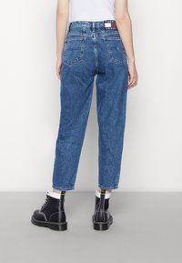 Osoba v modrých rovně střižených džínech s vysokým pasem a černobílých šněrovacích botách, stojící na pozadí jednoduchého bílého pozadí.