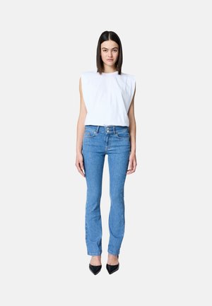 Femme aux cheveux raides et foncés portant un haut blanc sans manches avec épaules matelassées, un jean bleu taille haute et des chaussures noires à bout pointu, debout.