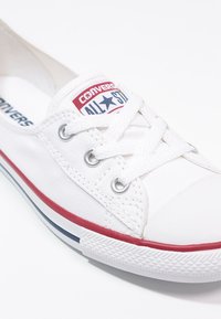 Białe trampki z czerwonymi i granatowymi akcentami, okrągły czubek i teksturowana gumowa podeszwa. Posiadają klasyczne sznurowanie i logo "Converse All Star".