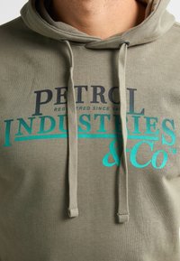 Felpa verde oliva con cappuccio e lacci, con stampa in blu e nero della scritta "PETROL INDUSTRIES & Co" in diverse dimensioni e stili di carattere.