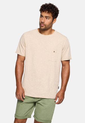 Hombre con cabello rizado que lleva una camiseta beige de manga corta con un pequeño bolsillo con botón y pantalones cortos verdes remangados, de pie contra un fondo blanco.