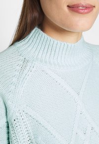 Ljusblå stickad tröja med hög ribbad krage, med texturerade mönster och en lös passform, som visar upp intrikata sömdetaljer.