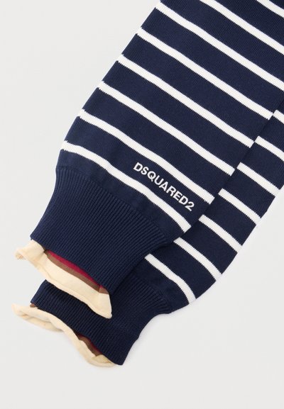 Μανίκια πλεκτού πουλόβερ σε ναυτικό μπλε χρώμα με λευκές οριζόντιες ρίγες και κεντημένο λογότυπο "DSQUARED2" κοντά στις ριμπ μανσέτες, στρωμένα πάνω από μπεζ ύφασμα με κόκκινες και καφέ ρίγες.