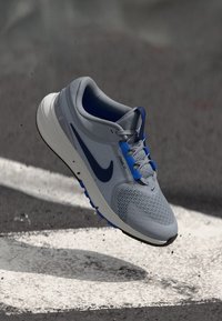 Adidași gri cu un superior din plasă și accente albastre, având o talpă intermediară căptușită și o talpă exterioară texturată pentru aderență.