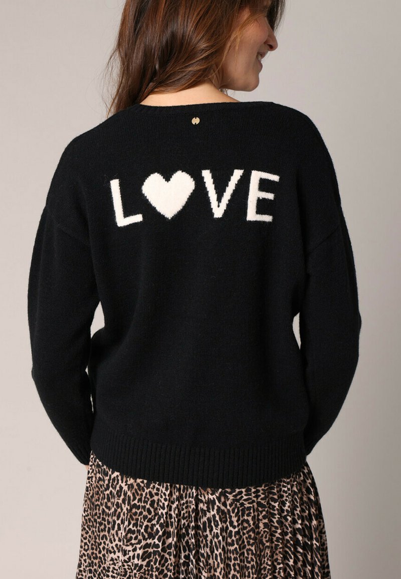 Pull noir avec l'inscription blanche "LOVE" et un design de cœur dans le dos. Texture douce, coupe oversize, ourlet côtelé, associé à une jupe à imprimé léopard.