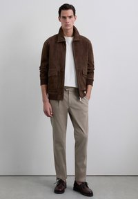 Chaqueta bomber de ante marrón con un gran cuello, dos bolsillos frontales, combinada con pantalones beige claros y zapatos marrones de cordones.