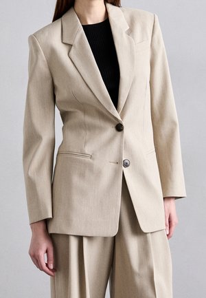 Beige räätälöity bleiser mustilla napeilla, päällä musta toppi ja beigeä vastaavat laskostetut housut seisovalla henkilöllä.