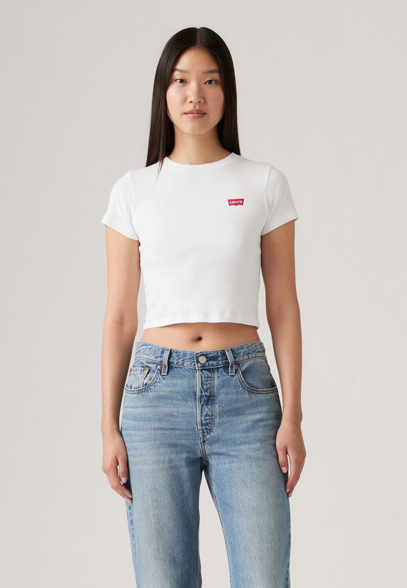 Νεαρή γυναίκα που φοράει ένα λευκό κοντό Levi's t-shirt και ψηλόμεσο μπλε τζιν, στέκεται μπροστά σε ένα απλό φωτεινό φόντο.