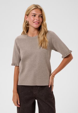 Femme blonde portant un pull taupe à manches courtes avec des poignets à volants, un pantalon noir, des boucles d'oreilles créoles dorées et des bracelets, souriante et regardant sur le côté.