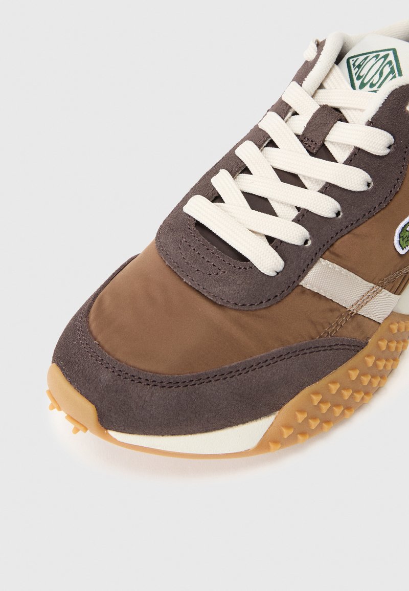 Zapatilla deportiva marrón que presenta una combinación de texturas lisas y de ante, cordones blancos y una suela de goma con un patrón y protuberancias.