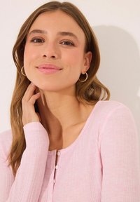 Light pink long-sleeve top with a ribbed texture, round neckline, and small buttons. Loose fit with fitted sleeves, and a soft fabric appearance.