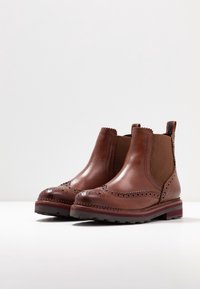 Hnědé kožené kotníkové boty s brogue detaily, elastickými bočními panely, vykrojenými okraji a silnou, texturovanou podrážkou s červeným pruhem.