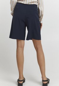 Námořnické modré šortky s hladkou texturou, délka po kolena a detailem zadní kapsy. Nosené s pruhovanou blůzou a černými sandály.