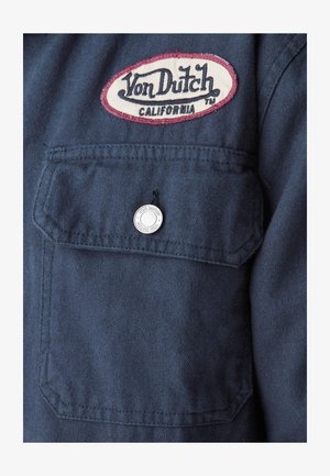 Patch logo Von Dutch California au-dessus d'une poche à rabat boutonnée sur un tissu texturé bleu foncé.