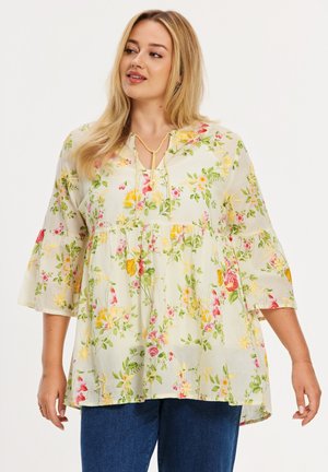 Femme blonde portant un chemisier floral ample avec manches cloche 3/4 et un jean bleu, regardant vers sa gauche avec un léger sourire.