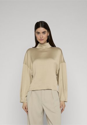 Femme aux cheveux noirs lisses portant un haut ample en satin beige à manches longues et un pantalon beige clair, debout devant un fond uni.
