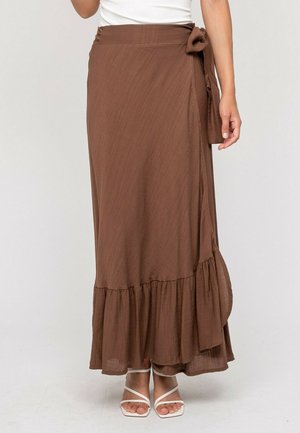 Jupe portefeuille marron, longueur cheville, avec ourlet à volants, nouée à la taille, portée avec des sandales blanches à brides et un haut blanc.